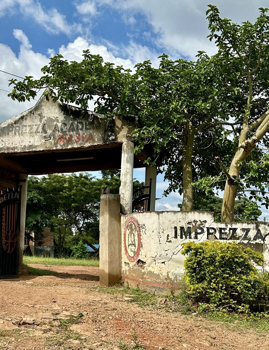 De poort van de Imprezza Academy in Busia, Kenia, met een verweerde muur en bomen.